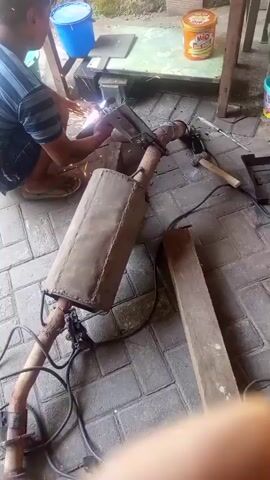A person wearing a striped shirt and shorts is kneeling on a paved surface, using an angle grinder on a metal object. Sparks are flying from the angle grinder. In the foreground, there is a large, rectangular muffler with a pipe attached. In the background, there is a wooden table with various items on it, including a blue bucket and a container of what appears to be food. The environment appears to be outdoors.