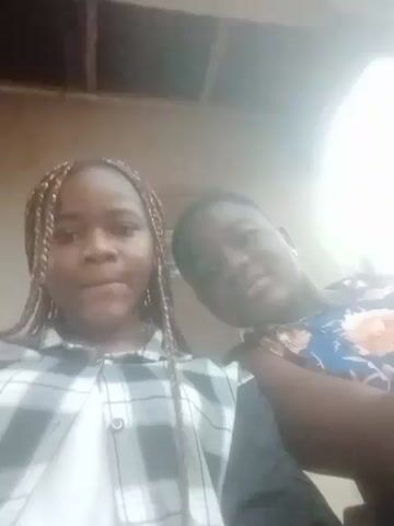 Two young women are posing close together for a selfie. The woman on the left has light brown braids and wears a white t-shirt under a plaid shirt. The woman on the right wears a floral-print dress. They are under an overhang or porch, with a door visible behind them. The lighting is bright and slightly overexposed.