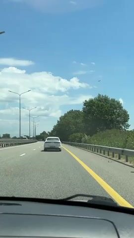 The video captures a first-person perspective from inside a vehicle driving on a multi-lane highway during the day. The sky is bright blue with scattered white clouds. Ahead, a white car travels in the same direction, staying within the lane marked by a solid yellow line on the right and dashed white lines on the left. To the right of the road, a dense line of green trees runs alongside a metal guardrail. On the left side, tall streetlights line the highway, and beyond them, what appears to be another lane or section of the road is visible with more distant vehicles, including a truck. A small, dark shape, possibly an airplane, is visible high in the sky in the distance. The overall scene depicts a calm, sunny day of travel on a well-maintained road. There is no visible text. The location context indicates Enem, Russia.