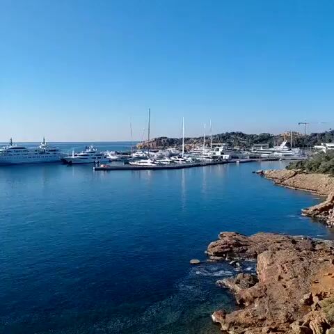 The video shows a view of a marina. Several yachts and sailboats are docked. The water is blue and the sky is clear. There are some rocks on the right side of the frame.