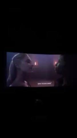 The video shows a darkened movie theater screen displaying a scene with two women. One woman on the left has pale skin and light blonde hair pulled back. The other woman on the right has dark skin and dark hair. Both women are facing each other and appear to be engaged in a conversation, as indicated by subtitles in Hebrew at the bottom of the screen. The background is dark with dim lighting suggesting an indoor setting. The video is dark and appears to be taken with a phone in a dimly lit theater, with the screen being the primary source of light.