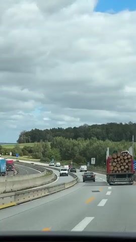 The video shows a view from a vehicle traveling on a highway. The highway has multiple lanes and is bordered by trees and vegetation. Several cars and a large truck carrying a load of logs are visible on the road. The truck is in the right lane and is the primary focus of the shot. The sky is cloudy.