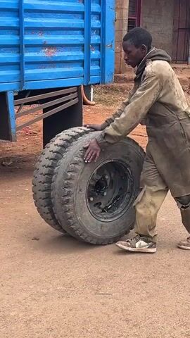 The video shows a person wearing dirty brown overalls pushing large truck tires. The person is standing next to a blue truck, and the tires are on the ground. The ground appears to be dirt or gravel. The person is pushing the tires with their hands. The tires are thick and have a tread pattern. The person's overalls are ripped in several places, particularly on the knees. The person is wearing sneakers. The truck has a blue bed and is parked on what appears to be an unpaved surface. The lighting suggests it is daytime, possibly overcast.
