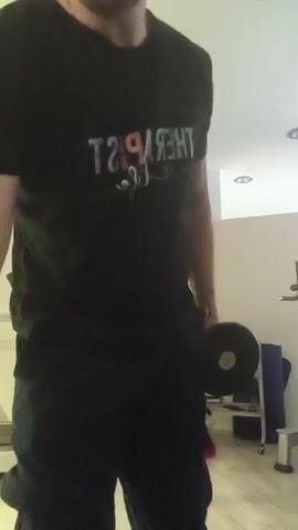 A man is performing bicep curls in a gym. He is wearing a black t-shirt with "THERAPIST LIFE" printed on it in colorful lettering. He is also wearing black cargo pants and white socks. The man is holding a dumbbell in each hand. In the background, there is a treadmill, a weight rack with more dumbbells, and a blue yoga mat. The room is well-lit with overhead lights. The man appears to be focused on his workout. There are no windows visible, so the time of day and weather are unknown. The presence of gym equipment indicates the location is a fitness facility.