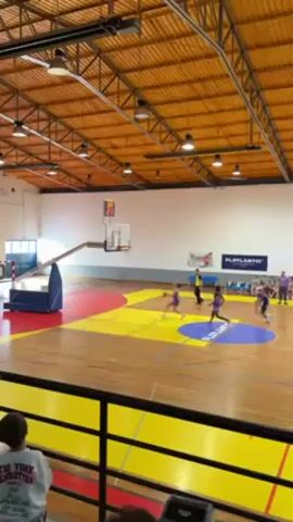 The video shows a basketball game taking place inside a gymnasium. Young girls wearing purple and white uniforms are playing on a basketball court with yellow, red, and blue painted sections. One of the girls shoots the ball and scores a basket. The court is made of wood and has basketball hoops at each end. The gym has a high ceiling with visible metal supports and light fixtures. Spectators are seated on benches along the side of the court. A referee in a black and yellow striped shirt is overseeing the game. A person wearing a gray sweatshirt with "NEW YORK MANHATTAN" printed on it is visible in the foreground, partially obscuring the view. Banners and advertisements are displayed on the walls.