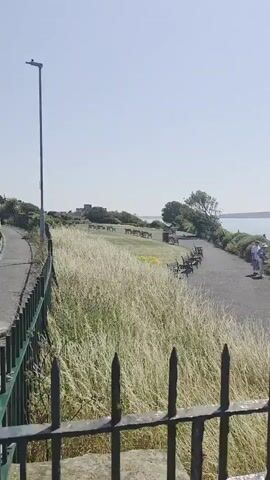 The video shows a scenic overlook on a sunny day. A paved path winds along a grassy slope, with benches placed along the side. In the distance, the sea stretches to the horizon, with a few boats visible. The video pans to reveal more of the coastline, including some buildings and a long pier extending into the water. A couple is seen walking along the path in the foreground.