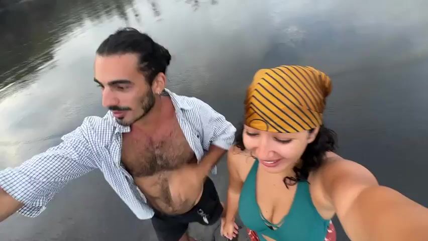 A man and a woman are standing close together in a body of water. The man is on the left, with his hair pulled back into a bun. He has a beard and mustache. He is wearing an unbuttoned checkered shirt and dark shorts. The woman is on the right, smiling, and has a yellow and black striped headscarf. She is wearing a green swimsuit top. The water is dark, with white foamy waves moving around them. The camera is close to their faces, and they are looking into the camera.