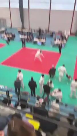 The video shows a judo match in a gymnasium. Two competitors wearing white judogi are engaged in a close combat on a red mat surrounded by green padding. Spectators are seated in the stands and other individuals in formal attire, presumably referees or officials, are situated at tables around the mat. A digital timer is visible on one of the tables. The gymnasium is large with a high ceiling and metal beams.