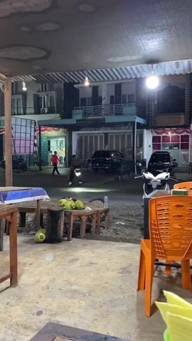 A motorcycle is ridden past a roadside stall at night. The stall is located in front of several multi-story buildings. The motorcycle is ridden by two people and is heading away from the camera. There are several coconuts on a table in the foreground. The ground is wet, suggesting it has recently rained. A bright light is visible in the background.