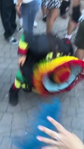 The video shows a person dancing on a brick paved surface in a crowded outdoor setting. The dancer is dressed in a black outfit with colorful stripes (green, yellow, red), dreadlocks, a mask, and a large, colorful hat. The person initially dances on the ground, spinning around, and then rises to dance upright. Surrounding the dancer are various people, some observing the dance and others walking by. The clothing of the observers varies; some are wearing jeans, dresses, shorts, and t-shirts. The environment appears to be a public space with brick paving stones.