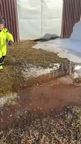The video depicts a toddler gleefully playing outside on a partly sunny day. The child, wearing a bright green and black snowsuit and a dark knit cap, is first seen splashing enthusiastically in a muddy puddle. The puddle is situated in an area of ground covered with patchy grass and some melting snow.  Behind the puddle is a barrier made of metal rods.  In the background there is a fence and a large pile of snow. The toddler then moves away from the puddle, walking toward a patch of ground still covered in ice and snow. The child smiles as they move.