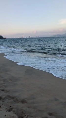The video shows a sandy beach with waves gently rolling onto the shore. In the distance, a city skyline is visible, punctuated by several high-rise buildings under a light blue sky with scattered clouds. The beach itself is a light tan color, marked with various footprints. The sea is a darker shade of blue, with white foam cresting the waves. There are no people visible in the video. The scene appears peaceful and serene.