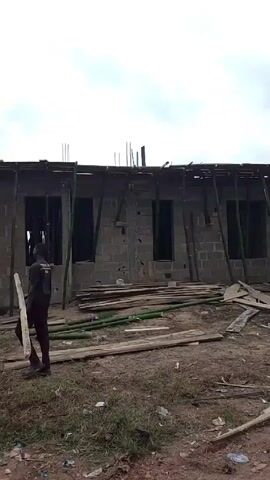 The video shows an unfinished one-story building under construction, viewed from ground level. The walls are built of grey cinder blocks, with window openings supported by green bamboo poles. The roof structure is in progress, with scaffolding and wooden planks in place. Two men are working on the roof, one of whom is handing down a wooden plank. There are scattered wooden planks and bamboo poles lying on the dirt ground around the building. The sky is overcast, and there is some vegetation visible on the ground. The environment appears to be a construction site in a somewhat rural or developing area.