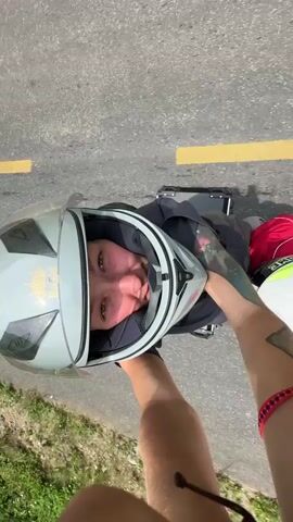 The video features two distinct clips, both from the perspective of someone riding a motorcycle.

The first clip (0:00-0:02) is a selfie-style shot, showing a person's arm holding the camera, pointed backward at two passengers. The main subject is a woman wearing a grey full-face motorcycle helmet, smiling at the camera. She has light-colored hair and is wearing a black top and red bottoms. Behind her, a second passenger, wearing a white helmet with green accents, is partially visible. A black top case is mounted on the motorcycle behind the woman. They are on an asphalt road with yellow dashed markings, under bright sunlight.

The second clip (0:02-0:05) switches to a forward-facing view, showcasing a scenic mountain road. The asphalt road, marked with yellow dashed lines, curves through a lush, mountainous landscape. On the left, a steep, rocky mountain face is covered with green vegetation. On the right, the terrain drops away, revealing distant mountains under a partly cloudy sky. Utility poles and power lines run along the side of the road. Below the road level, some small, simple buildings, likely houses or shacks, with corrugated tin roofs are visible. The setting is a rural, mountainous area, consistent with Minh Sơn Commune, Vietnam, under clear daylight conditions.