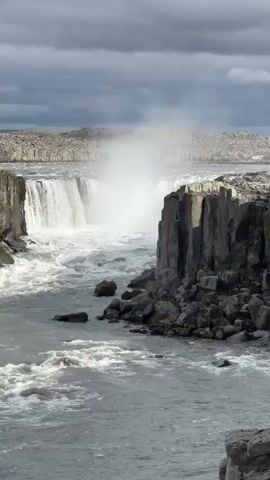 The video captures a dramatic view of a powerful waterfall and the turbulent river it feeds. A vast curtain of milky grey water plunges over a wide, dark cliff face, generating immense spray and mist that rises high into the air. The waterfall is flanked by steep, rugged cliffs composed of dark, possibly columnar basalt rock formations. The river below churns violently with white rapids and swirls as it flows downstream, carrying large rocks. The surrounding landscape beyond the immediate cliffs appears barren and rocky. The sky is overcast with heavy, dark grey clouds, lending a dramatic and somewhat subdued light to the scene. No people or visible text are present. The footage highlights the raw power and grandeur of nature, consistent with Iceland's volcanic and glacial landscapes.