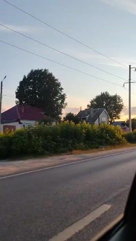 The video captures a view from a moving vehicle on a paved road. The scene is a rural or suburban area with houses and trees visible on the side of the road. Power lines run overhead. The sun appears to be setting, casting a warm light on the scene and creating a soft glow on the houses and foliage. A dark-colored sedan is seen driving in the opposite direction.