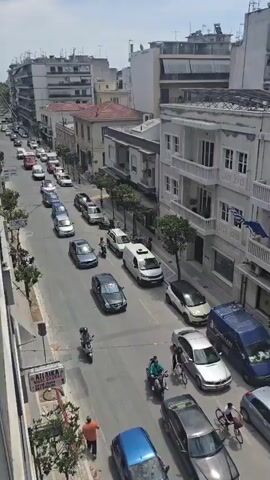 The video shows a busy street scene from an elevated perspective. Cars, scooters, and pedestrians move along the road and sidewalks.  Buildings line both sides of the street, with a mix of architectural styles.  A sign with a large yellow "E" is visible in the foreground.  Further down the street, the view extends to more buildings and cars, with some construction work taking place in the distance. The overall scene depicts a typical urban environment with everyday activities.