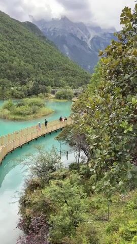 The video displays a scenic landscape with a turquoise lake bordered by lush green mountains and a snow-capped peak in the distance. A wooden bridge, curved in shape, spans across the water, connecting two landmasses. Several people are seen walking on the bridge. The bridge is made of wooden planks and features railings on both sides. Some individuals are walking towards the right side of the bridge, while others are moving towards the left. The water is clear and reflects the surrounding greenery and sky. Small islands covered in vegetation are scattered within the lake. The overall environment appears to be a natural park or a tourist destination, with the bridge serving as a pathway for visitors to enjoy the view. The lighting suggests it is daytime with some cloud cover.