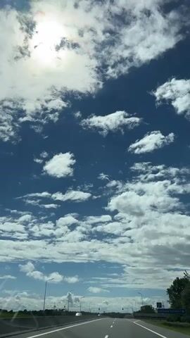 A view from a vehicle traveling on a highway on a bright, cloudy day. The sun is partially visible through the clouds. The road is a multi-lane highway with white lane markings. There are streetlights and signs visible along the side of the road. The sky is a deep blue with scattered white clouds.
