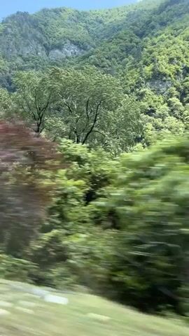 The video shows a view from a moving vehicle, likely a train, looking out at a lush green mountainside. The camera captures a dynamic scene with trees and vegetation blurring as the vehicle speeds past. In the background, a steep, rocky mountain face is visible, partially covered in green foliage. The sky above is a mix of blue with scattered white clouds. A notable feature on the mountainside appears to be a man-made structure, possibly a road or a building, with a long, white concrete element running along it. The overall impression is one of a scenic journey through a natural, mountainous landscape.