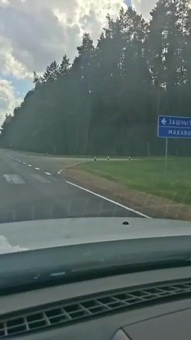 The video is filmed from the perspective of someone driving a vehicle. The road ahead curves to the right, bordered by trees on the left and grass on the right. The sky is partly cloudy. As the video progresses, the road straightens and continues through a landscape of fields and trees under a blue, cloudy sky. The dashboard of the vehicle is visible in the foreground.