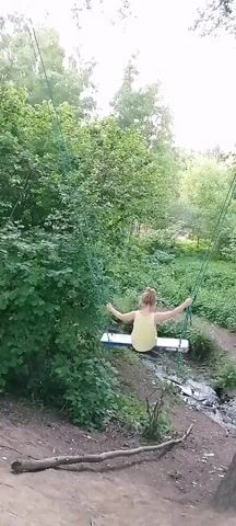 The video shows a young girl with blonde hair in a ponytail, wearing a yellow dress, swinging on a handmade swing set in a wooded area. The swing has a wooden seat and is suspended by green ropes from a higher structure, likely a branch or an overhead support. The girl is actively swinging, with her arms extended to the sides. The background is filled with lush green trees and foliage, suggesting a natural, outdoor environment. A small stream or ditch runs alongside the path below the swing. The ground is covered in dirt and fallen leaves, with a few branches scattered on the path. The lighting appears to be natural daylight, casting soft shadows.