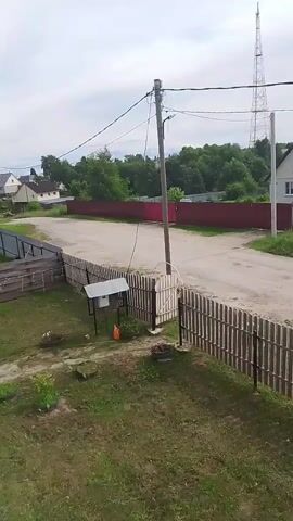 The video shows a panning view of a rural landscape. A wooden fence is in the foreground, with a grassy yard behind it. A paved road runs horizontally across the mid-ground, bordered by another fence. On the other side of the road, a house with a red roof is visible, along with trees and a tall radio tower in the distance. The sky is overcast.