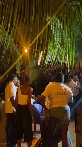 A group of young adults, mostly wearing light-colored tops, are dancing and cheering under palm trees at night. A string of lights and a single lamp illuminate the scene. Some people are clapping, while others are jumping and raising their arms. One person in the center of the frame is wearing a yellow shirt with the letters “ESAE” printed on the back. The ground appears to be sandy. A few blue plastic chairs are scattered around the edge of the group. Some people are smiling and laughing. The overall atmosphere is energetic and celebratory.