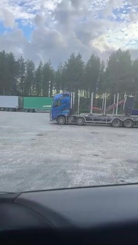 The video shows a blue logging truck parked in a gravel lot, with a line of trees in the background. Another truck, a white and red semi-trailer, is parked to the left of the logging truck. The sky is cloudy. The camera appears to be positioned inside a vehicle, looking out through the windshield.
