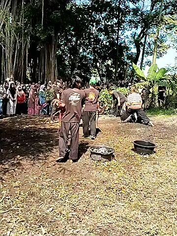 A martial arts demonstration occurs outdoors on a dirt patch covered in dry leaves and grass. A crowd of people of various ages and ethnicities watches the performance from behind a row of trees. Several men in black pants and dark t-shirts participate, some with green hats. One man swings a whip. Three black containers of some sort sit in a line on the ground. Several other men kneel down on the ground. Trees are visible in the background. The sun shines brightly overhead.
