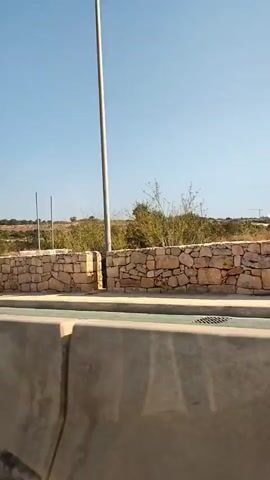 The video shows a low-angle, side-view of a moving vehicle, likely a car, passing by a rural landscape on a bright, sunny day. The foreground is dominated by the concrete barrier of the road. Beyond the barrier, a low, dry-stone wall made of rough, light-colored stones runs horizontally across the frame. Behind the wall, the terrain is uneven and covered with sparse, dry vegetation, including bushes and possibly some low trees. The sky above is clear and a vibrant blue. There are no people visible in the video, and the primary action is the movement of the vehicle, causing the scenery to scroll by. A thin, vertical pole with some equipment attached to the top can be seen near the right side of the wall. The overall impression is one of a journey through a somewhat arid, natural environment.