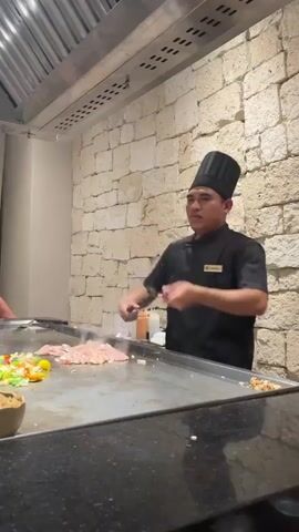 A male chef wearing a black uniform and chef hat stands behind a teppanyaki grill. He is using two knives to chop food on the grill. The grill has raw chicken and vegetables on it. The background is a stone wall.