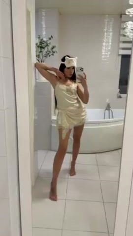 A woman is taking a selfie in a bathroom. The bathroom is tiled white and contains a jacuzzi bathtub. She's wearing a satin nightgown with lace trim and a sleep mask with bear ears. She leans against the wall and waves.