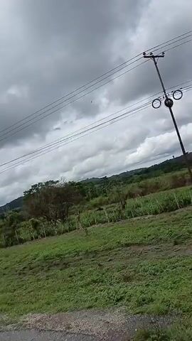 The video shows a view from a car driving on a road. The scenery includes green fields, trees, and a cloudy sky. There are power lines overhead. The car is moving, as evidenced by the changing scenery and the presence of the side mirror in the shot. The weather appears overcast.