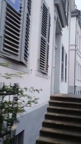 The video begins by showing the exterior of a white building with closed shutters. Steps lead up to the building's entrance. The camera then pans to the right, revealing a cobblestone street lined with buildings and outdoor seating areas for cafes or restaurants. Several people are walking along the street, including a couple holding hands. Bicycles are parked along the side of the street. In the background, a construction crane is visible. The sky is overcast.