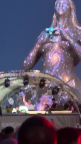 The video shows a nighttime outdoor event with a large statue in the background. The statue is projected with swirling, starry patterns resembling a galaxy. In the foreground, there is a stage with a DJ booth. A DJ wearing a white shirt and glasses is visible, raising his hand and smiling. Other individuals are present on the stage, likely part of the DJ crew. In front of the stage, a crowd of people is gathered, with some raising their hands. Red and white lights are visible in the lower part of the frame, illuminating the audience. The overall atmosphere appears to be that of a music festival or concert. The text "EXIT2" is visible on a banner in front of the stage.