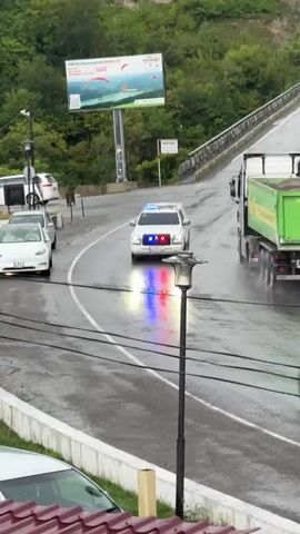 A white SUV with flashing blue and red lights on its grille is driving down a wet, winding road. The SUV has "LIBERTY" written on its side. There are other vehicles on the road including a green dump truck and a white van. A billboard is visible in the background. The road is lined with trees and a rocky incline. There is a parking sign on the right side of the road.