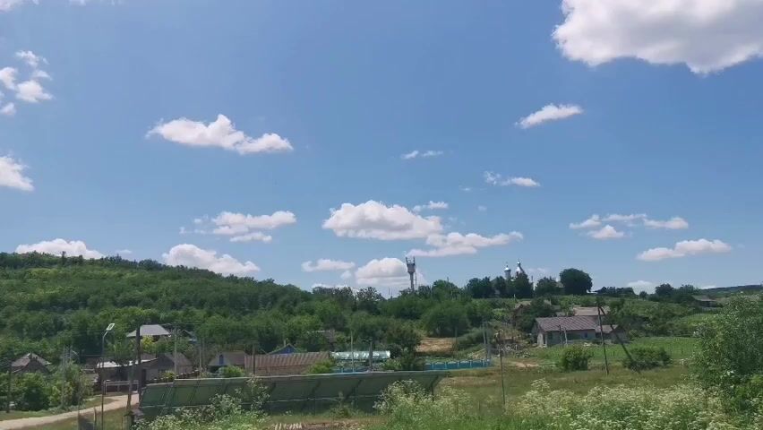A wide shot captures a rural landscape under a clear blue sky dotted with white clouds. In the foreground, solar panels are visible, positioned on the ground and angled upwards. A dirt road or path runs diagonally through the scene. Further into the distance, a small village is nestled among rolling green hills covered in trees. Several houses with roofs of various colors are scattered throughout the village. A tall communication tower rises above the trees on one of the hills. The overall impression is one of a peaceful, sun-drenched countryside.