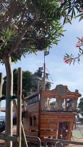 The video shows a child playing on a wooden pirate ship structure in a playground. The structure is designed to resemble the stern of a pirate ship, complete with a mast, black sails, and a pirate flag flying from the top. The child is wearing a white shirt and yellow shorts and is seen walking around on the deck of the ship. The playground is outdoors and surrounded by trees. The sky is blue with some white clouds.