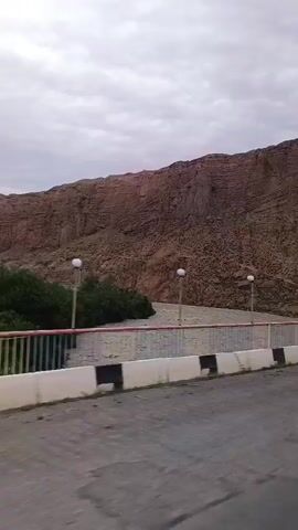 The video shows a roadway with a mountain in the background. There is a concrete barrier with black and white stripes along the side of the road. Several streetlights are visible, one of which has a flag attached to it. The sky is overcast.