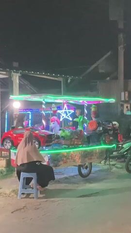 A nighttime scene shows a mobile carousel with several children riding in toy vehicles. The carousel is decorated with colorful lights, including green lights outlining the platform and a star in the center. There are also other decorative elements like what appear to be ice cream cone shapes. One person, wearing a hijab, sits on a blue stool watching the carousel. In the foreground, a person on a motorcycle is blurred due to motion. The background suggests an outdoor setting, possibly a street or market, with buildings and other vehicles visible.