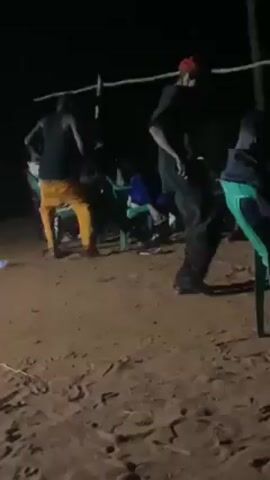 At night, several people are congregated on a patch of ground with sparse vegetation.  Some individuals are standing and dancing, while others are seated on plastic chairs scattered around. The lighting is dim, and a structure made of slender poles stands overhead. The ground appears to be sandy or dusty. The people are wearing a variety of casual clothing, including a red baseball cap.