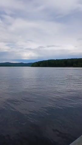 The video displays a body of water, likely a lake, under a cloudy sky. The water is dark and appears calm with small ripples. In the distance, across the water, a tree-lined shore can be seen, with hills behind it. There is a small, light-colored object, possibly a boat or kayak, visible on the water in the middle distance. The overall scene is peaceful and natural.