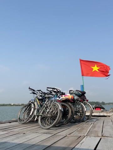 The video captures a serene daytime scene on what appears to be a boat or a wooden pier. In the foreground, several bicycles are parked closely together on weathered wooden planks, facing towards the right side of the frame. Some of the bicycles have luggage or backpacks strapped to their rear racks, suggesting they are part of a tour or travel.

Dominating the mid-ground is a vibrant red flag with a large yellow star, the national flag of Vietnam, fluttering in the wind from a pole. The flag's fabric shows some wear and tear.

Beyond the wooden platform and the flag, a calm body of water, likely a river or a large canal, stretches into the distance. The far bank is lined with lush green trees and vegetation under a clear, bright blue sky with a few wispy white clouds.

The audio accompanying the video features the distinct chugging sound of a boat engine, indicating that the bicycles are on a moving vessel. Faint, indistinct human voices can also be heard in the background. The overall atmosphere suggests a peaceful journey by water in a scenic, possibly rural, part of Vietnam.