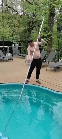 A man is shown using a pool vacuum to clean a swimming pool. He is fair-skinned, dark-haired, and is wearing black pants and brown slip-on shoes. He is shirtless. The swimming pool is blue. The surrounding deck is wooden. Several lounge chairs are placed around the deck. Trees are visible in the background. The man is moving the pool vacuum back and forth in the pool.