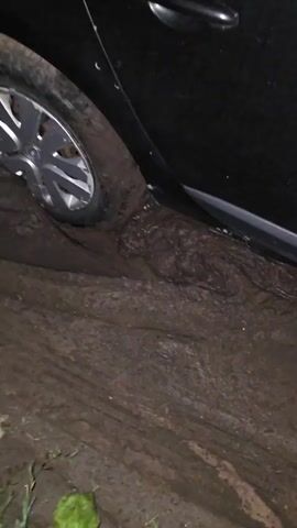 A black car is stuck in deep mud. The front passenger-side tire is submerged in the mud, with the vehicle's side panel resting on the muddy ground. The mud is thick and viscous, showing deep tire tracks. The surrounding environment is dark and appears to be a dirt road or unpaved area. Green vegetation is visible along the edge of the mud.
