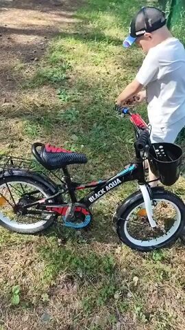 A young boy wearing a white t-shirt and a black baseball cap with a yellow emblem stands next to a black and red bicycle. The bicycle has a basket on the front, a black chain guard with a blue design, and a red and black seat. The boy appears to be interacting with the bicycle, possibly adjusting it or preparing to ride it. He is wearing a white t-shirt and a black baseball cap with a yellow emblem that reads "RUSSIA". The scene is outdoors, with a grassy area and trees in the background, suggesting a park or a wooded area. A green metal fence runs along the right side of the frame. The lighting suggests it is daytime, with sunlight filtering through the trees. The boy's posture and interaction with the bike suggest he is engaged and possibly excited. The text "BLACK AQUA" is visible on the frame of the bicycle.