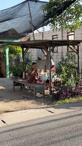 The video opens with a street-level view of an outdoor seating area under a makeshift shelter. Two people are present. A man in a patterned shirt and red pants sits on a wooden bench, looking down at something on a tray before him. To his right, a woman wearing a grey hijab and a patterned top is also seated, seemingly engaged in a task with her hands over a white bowl.

The shelter is constructed from wood and covered with a black mesh tarp, providing shade. In front of the shelter, there's an abundance of plants, with lush green foliage and vibrant purple bushes in the foreground. Beyond the shelter, a building with cinder block walls is visible. The overall environment suggests a warm, outdoor setting, possibly a residential area or a small business. The time of day appears to be daytime, with sunlight casting shadows.