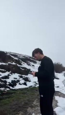 A man stands outside on a partly snowy day looking at his phone. He has brown hair and is wearing a black sweatshirt and black sweatpants. He says, "These guy are crazy." The area around him is dirt and patches of snow. Beyond him is a muddy hill with patches of snow. The sky is white.