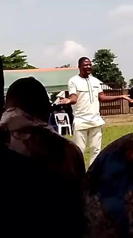 A man in a light-colored outfit is speaking outdoors, gesturing with his hands. He is standing in front of a group of people, some of whom are seated on chairs. A tent structure and some trees are visible in the background. The weather appears to be sunny.